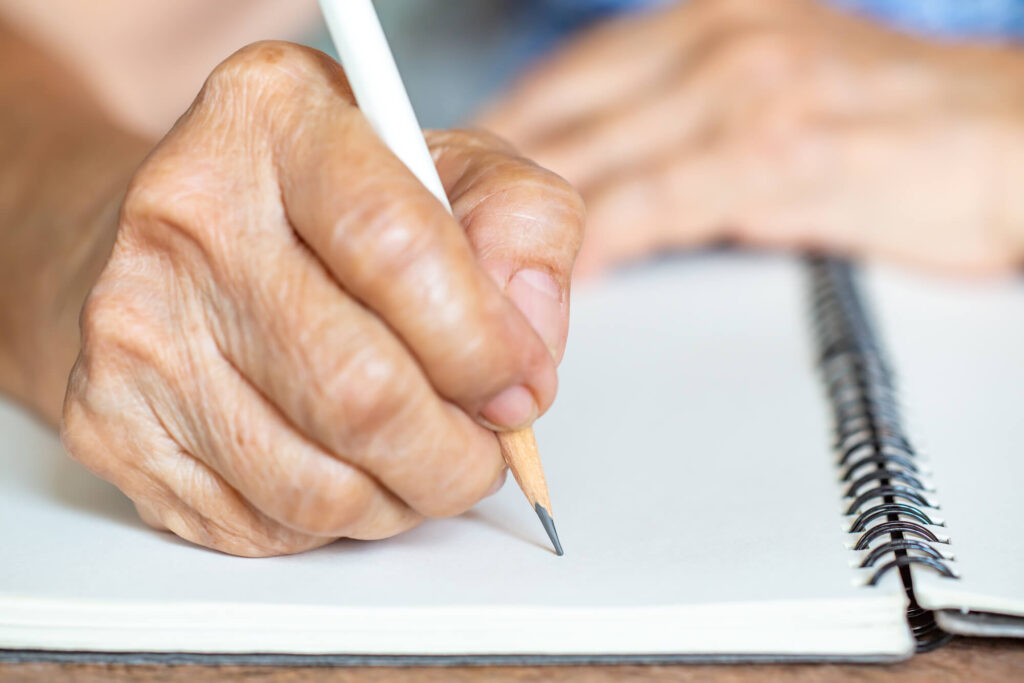 Eine ältere Dame sitzt an einem Tisch und schreibt mit einem Stift auf ein Blatt Papier.